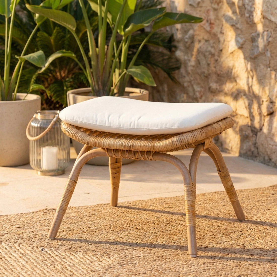 Wicker stool with a white cushion on a textured surface with plants and a stone wall in the background.