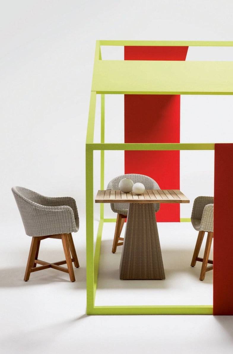 Square single-leg dining table from the Gipsy Collection with woven pedestal base and wooden top, shown in a styled hospitality interior setting.